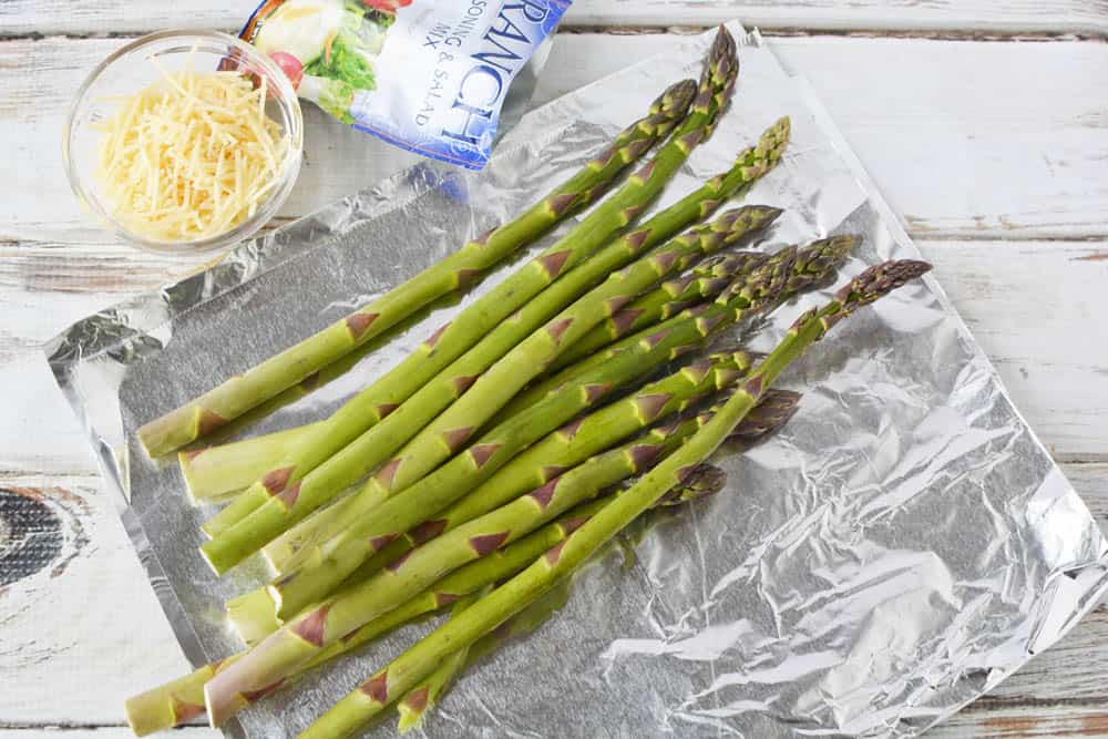 Fresh asparagus on a piece of foil with a ranch dressing packet mix and small bowl of Parmesan cheese.