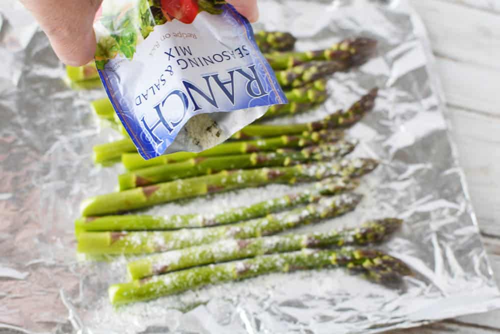 A packet of ranch dressing mix being poured over asparagus on foil