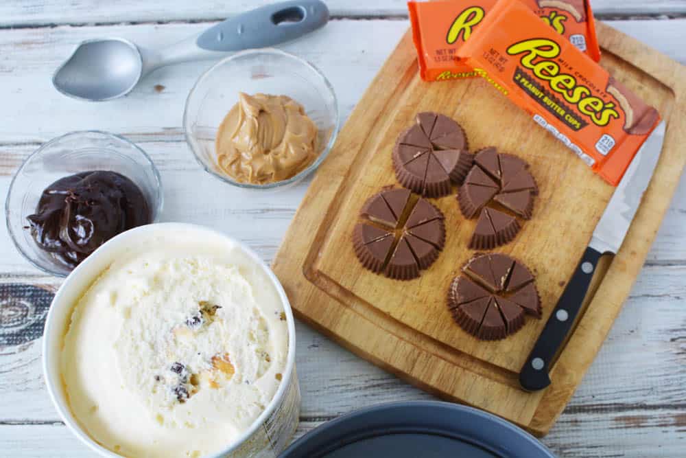 A tub of ice cream with bowls of fudge and peanut butter next to a cutting board with cut up Reese's Peanut Butter cups.