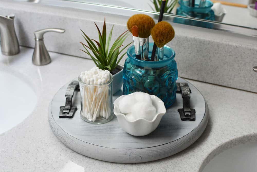 Round wood tray on bathroom counter with brushes, cotton balls, cotton swabs and a plant