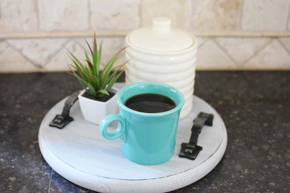 Turquoise coffee mug filled with coffee on a tray with a plant and white canister on counter